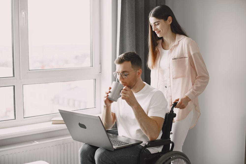 A man in a wheelchair with a laptop and a coffee, a support worker behind him