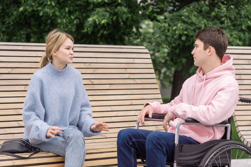 A support worker and a young man in a wheelchair talking on a park bench